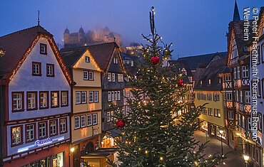Weihnachtlicher Marktplatz und Burg im Nebel Beleuchtete Fachwerkhäuser und ein geschmückter Weihnachtsbaum in einer abendlichen Stadtansicht.