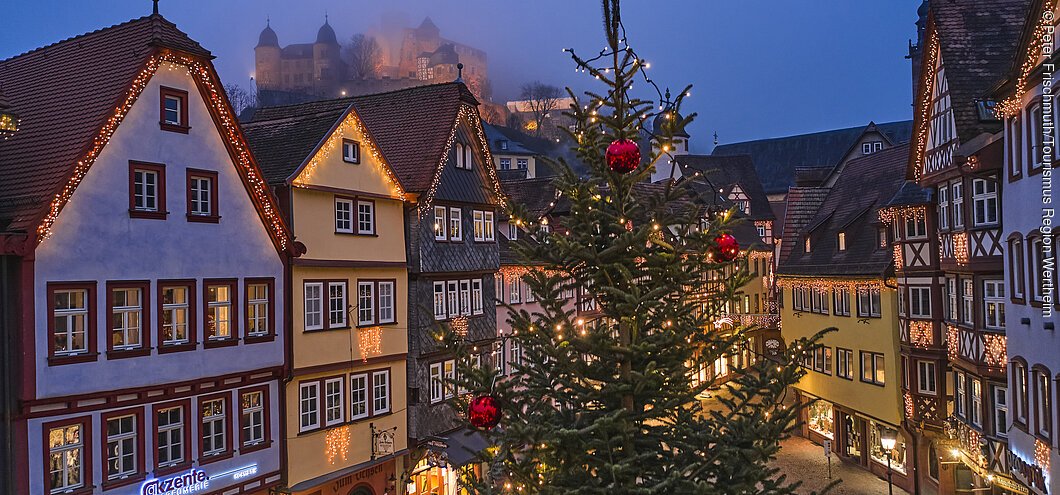 Beleuchtete Fachwerkhäuser und ein geschmückter Weihnachtsbaum in einer abendlichen Stadtansicht.