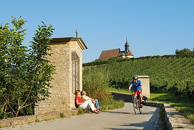 Zwei Personen sitzen an einer Steinmauer, ein Radfahrer f&auml;hrt auf einem Weg neben Weinbergen und einer Kirche.